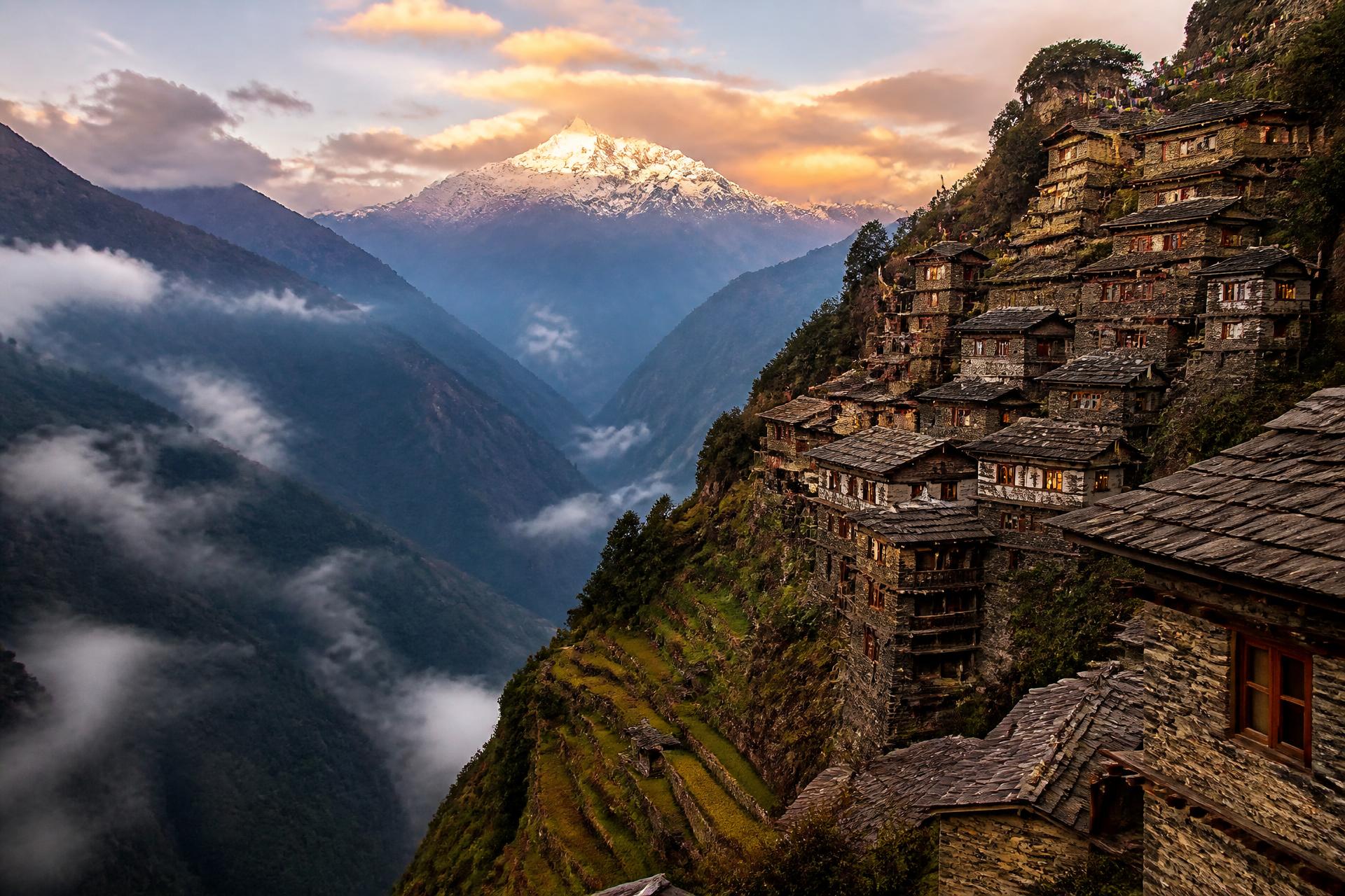 Himalayan cliffside village at sunset — ancient stone houses cascading down a mountainside, terraced fields, prayer flags, snow-capped peak glowing above misty valleys
