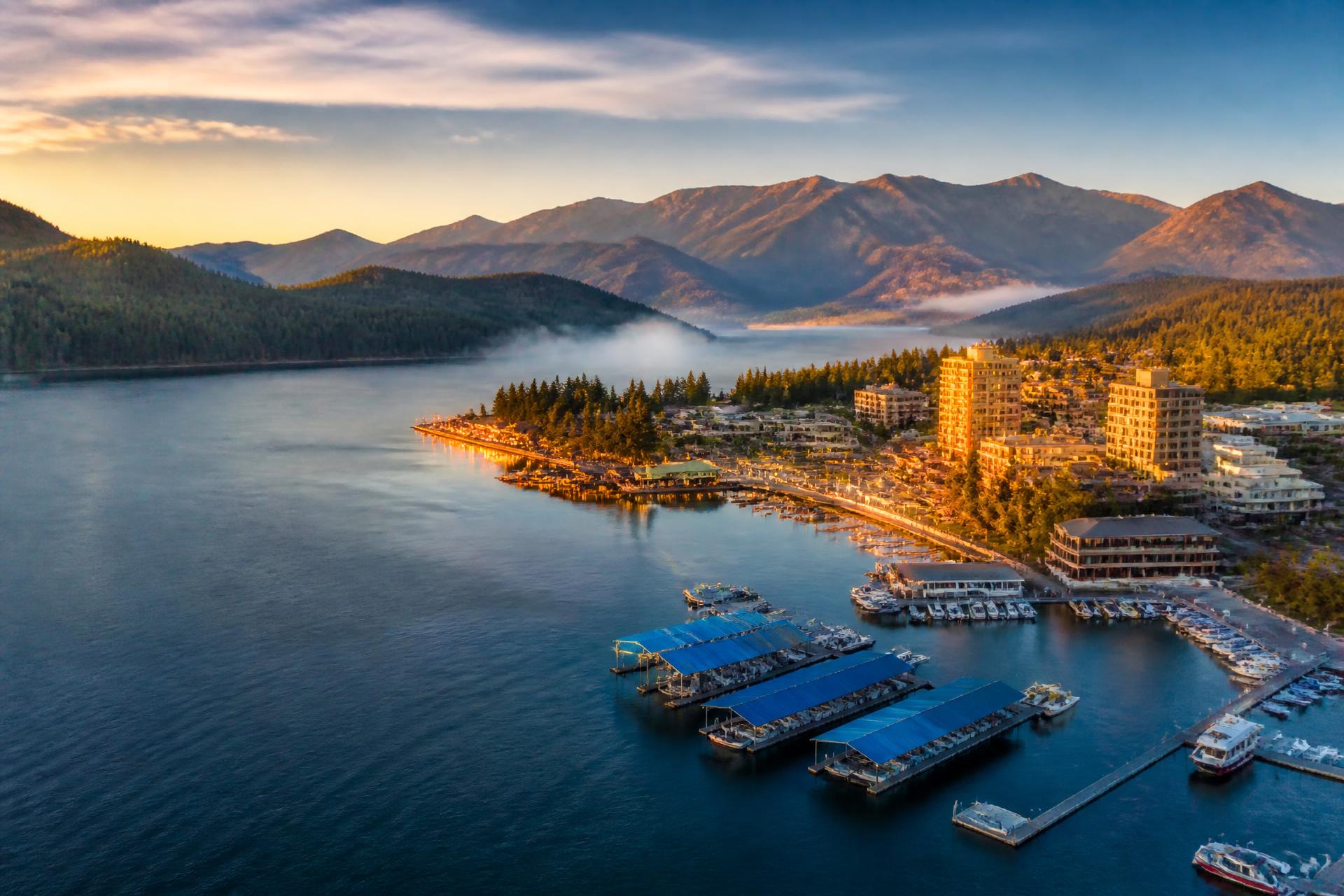 Coeur d'Alene, Idaho at golden hour — the sparkling lake, waterfront docks, and forested mountains of the American Northwest glowing under a warm sunset